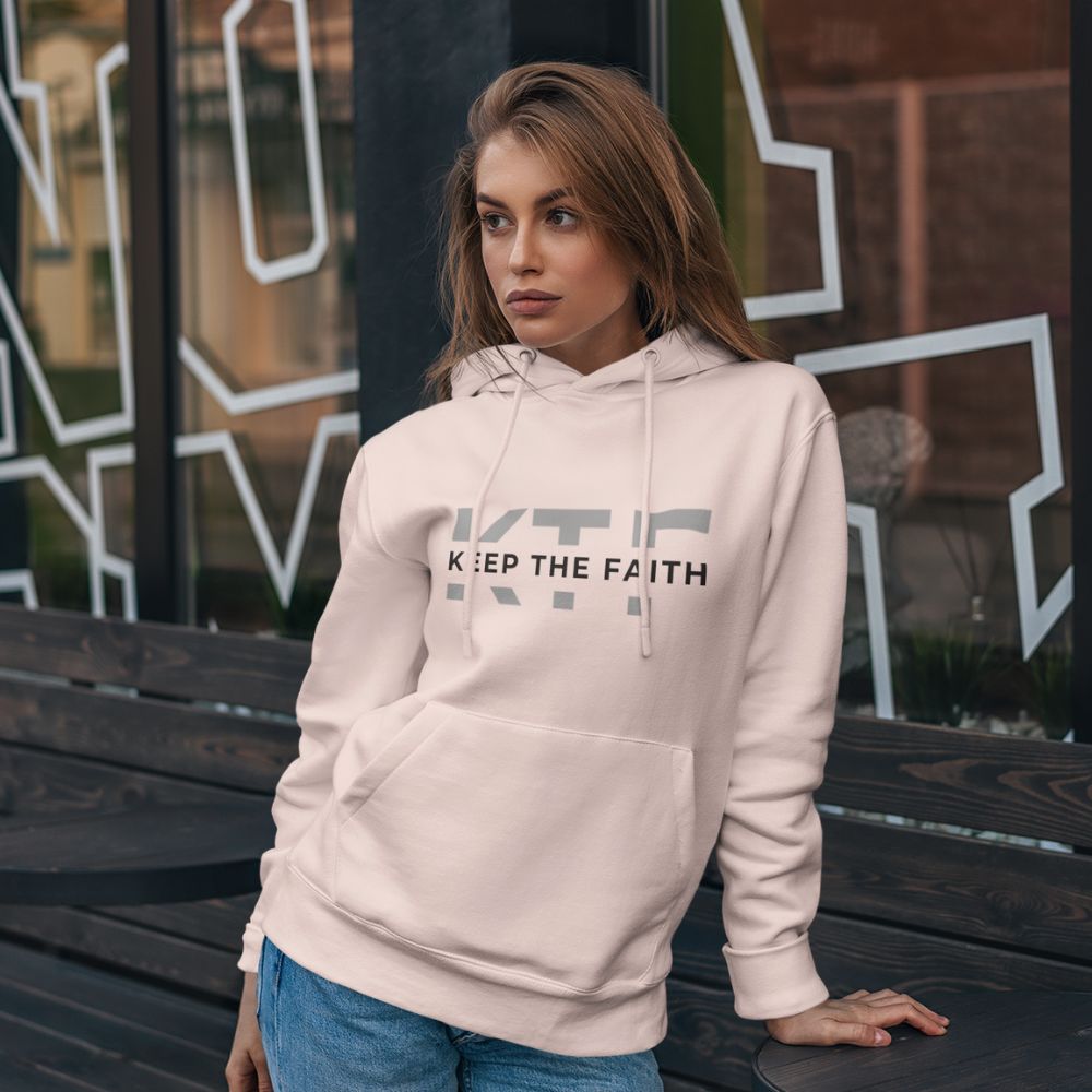 Woman wearing light pink Keep The Faith hoodie sitting outdoors by modern building
