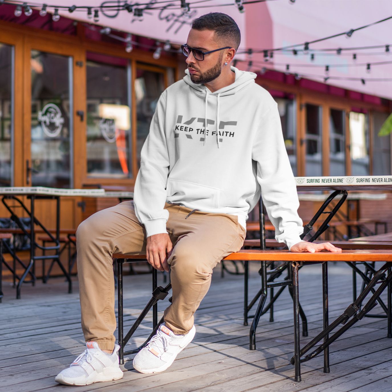 Man wearing light Grey Keep The Faith hoodie sitting outdoors by modern Restaurant 